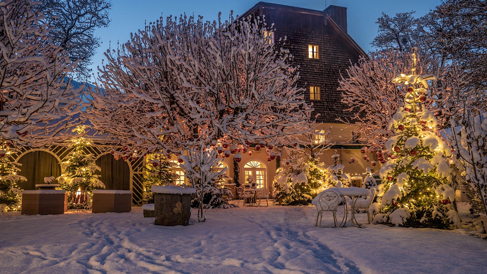 Das Bild zeigt die elegante Gartenterrasse in der Abenddämmerung. Ein Steinweg schlängelt sich durch den gepflegten Rasen, gesäumt von bequemen Loungemöbeln. Gäste speisen unter großen weißen Sonnenschirmen, umgeben von üppigem Grün und alten Bäumen. Das historische Gebäude im Hintergrund, teilweise mit Efeu bewachsen, verleiht der Szenerie besonderen Charme. Warmes Licht aus dem Inneren und von der Terrasse schafft eine gemütliche, einladende Atmosphäre. Die Szene strahlt Ruhe, Eleganz und eine perfekte Verbindung von Natur und gehobener Gastlichkeit aus.