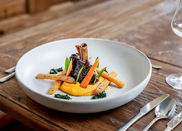 Dieses Gericht präsentiert sich als moderne Interpretation eines klassischen Tomaten-Mozzarella-Salats. Verschiedene bunte Tomatensorten, darunter Cherry- und grüne Zebra-Tomaten, sind kunstvoll mit cremiger Avocado-Creme angerichtet. In der Mitte thront ein zartes Stück Büffelmozzarella, bestreut mit frisch gemahlenem Pfeffer oder Gewürzen. Frisée-Salat sorgt für Frische und Struktur. Ein aromatisches Dressing rundet das Gericht geschmacklich ab. Die Präsentation auf einem schlichten weißen Teller betont die leuchtenden Farben und die sorgfältige Anordnung der Zutaten – ein eleganter, leichter und geschmackvoller Einstieg in ein Menü oder als raffinierter Hauptgang.