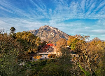 This stylishly furnished bedroom radiates warmth and elegance. The combination of green-painted walls, a classic wooden bed, and herringbone parquet flooring creates a harmonious atmosphere. Two bedside tables with decorative lamps flank the inviting bed, adorned with white linens and patterned accent pillows. Light curtains frame the windows, allowing plenty of natural light to fill the space. A desk with fresh flowers and a water bottle, along with a comfortable armchair, completes the setting. The soft lighting and use of natural materials contribute to a calm and cozy ambiance, ideal for relaxation.