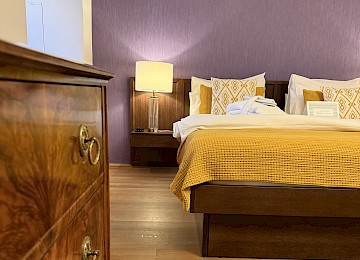 This stylish bedroom blends classic elegance with cozy comfort. A large wooden bed with white linens, decorative throw pillows, and a soft blanket serves as the room’s focal point. The green-painted wall and herringbone parquet flooring add warmth and character. Matching bedside tables with lamps provide soft ambient lighting. Large windows with light curtains let in plenty of natural daylight. An antique desk with fresh flowers and a comfortable chair complete the harmonious setup. The combination of natural materials and soft colors creates a calm, inviting atmosphere—perfect for rest and relaxation.