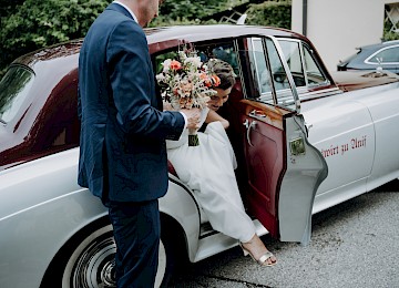 Rolls Royce (c) Gassner Gastronomie: The image shows an elegant silver vintage Rolls-Royce, beautifully decorated for a wedding. White ribbons and floral arrangements adorn the door handles and hood, giving the car a festive and romantic touch. The gleaming chrome grille and classic design highlight the vehicle’s luxurious character. In the background, there