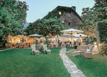 Dieses stimmungsvolle Bild zeigt einen idyllischen Garten eines Restaurants oder Landhotels in der Abenddämmerung. Ein gepflasterter Weg führt durch den gepflegten Rasen zu gemütlich beleuchteten Sitzbereichen unter weißen Sonnenschirmen. Gäste genießen entspannt ihr Essen oder Drinks im Freien, umgeben von üppigem Grün, blühenden Pflanzen und alten Bäumen. Bequeme Liegestühle und Loungemöbel laden zum Verweilen ein. Das historische Gebäude im Hintergrund mit bewachsener Fassade verleiht dem Ambiente besonderen Charme. Die harmonische Beleuchtung und das natürliche Setting schaffen eine warme, einladende Atmosphäre – ideal für einen Sommerabend in geselliger Runde.