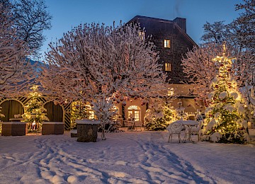 Diese stimmungsvolle Winteraufnahme zeigt einen festlich geschmückten Hof bei Nacht. Die Bäume sind dick mit Schnee bedeckt und mit leuchtenden Lichtern sowie roten Christbaumkugeln dekoriert, die eine warme, märchenhafte Atmosphäre erzeugen. Im Hintergrund erhebt sich ein charmantes, altes Gebäude mit Holzfassade, dessen Fenster von innen hell erleuchtet sind. Eine weiße Schneedecke überzieht den gesamten Hof und reflektiert das warme Licht der Dekoration. Die Szene strahlt Ruhe, Besinnlichkeit und winterliche Festlichkeit aus – ein perfekter Ort für ein stimmungsvolles Weihnachtsfest oder romantische Winterabende.