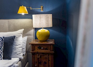 This image shows a beautifully detailed bed in a cozy hotel room. The focus is on decorative cushions in mustard yellow and white, featuring a geometric pattern that perfectly matches the mustard yellow bed throw. The padded gray headboard adds an elegant touch to the setting. To the left, a wooden nightstand holds a stylish yellow lamp that casts a warm, soothing light. The combination of warm tones, soft textures, and clean lines creates an inviting and modern atmosphere—ideal for restful nights in a space that blends comfort with style.