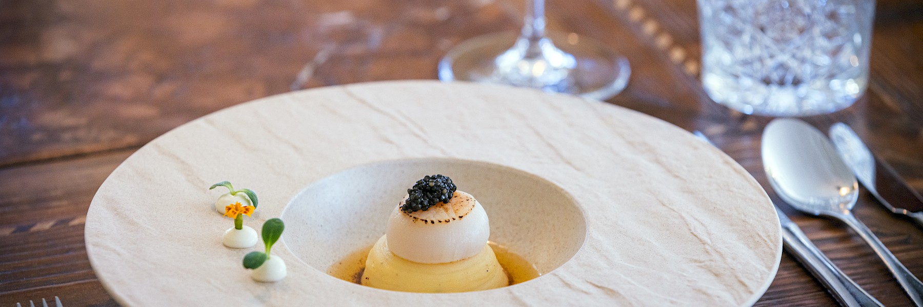 Das Bild zeigt ein elegant angerichtetes Gourmetgericht in einer rustikalen, hellen Keramikschale auf einem Holztisch. Im Zentrum liegt ein perfekt gebratenes Fischfilet mit knuspriger Haut, umgeben von hausgemachter Pasta, grünem Spargel und fein geschnittenem Zucchini. Kleine Blüten und zarte Kräuter sorgen für dekorative Akzente. Eine helle Schaumsoße ergänzt das Gericht optisch und geschmacklich. Die Präsentation wirkt modern und gleichzeitig naturverbunden – ein kunstvoll inszeniertes Gericht, das Frische, Qualität und Raffinesse ausstrahlt. Ideal für ein gehobenes kulinarisches Erlebnis.