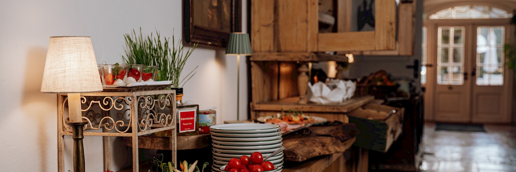 (c) Gassner Gastronomie: The image shows a beautifully arranged buffet in a charming, rustic interior. A long wooden counter displays bowls of fresh vegetables like radishes, cherry tomatoes, and carrots, alongside a vibrant platter of colorful raw vegetable salad. Small glasses with appetizers, potted herbs, a selection of oils, and a neatly stacked pile of plates complete the setup. Soft lamp lighting creates a warm and inviting atmosphere. In the background, there are bread baskets, cold cuts, and double doors leading to a bright outdoor area. The scene feels high-quality and welcoming—ideal for a cozy breakfast or brunch buffet.