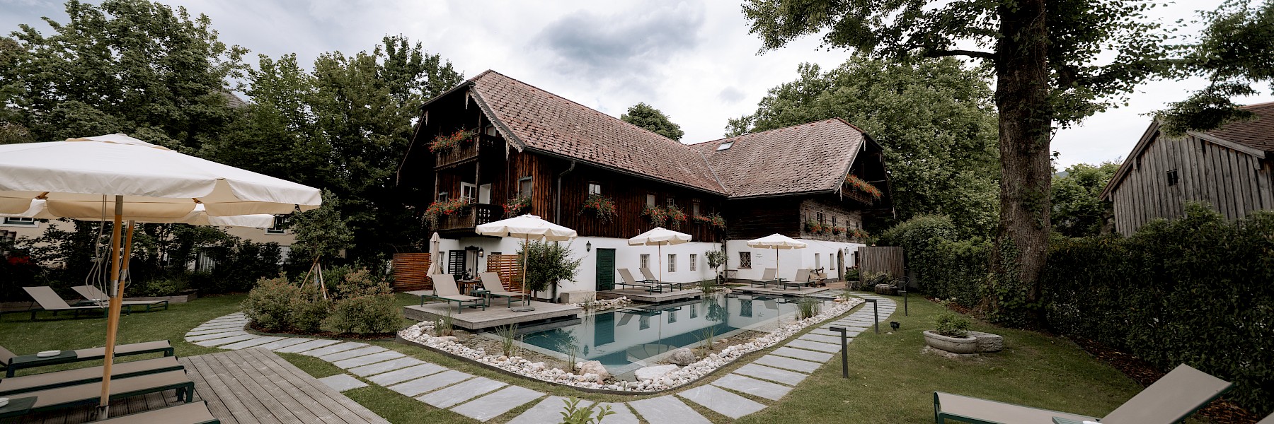 (c) Hechtfisch: Das Bild zeigt einen idyllischen, gepflegten Garten mit einem Naturpool, umgeben von Liegestühlen und Sonnenschirmen vor einem charmanten, alten Landhaus mit Holzbalkon und blumengeschmückten Fenstern.