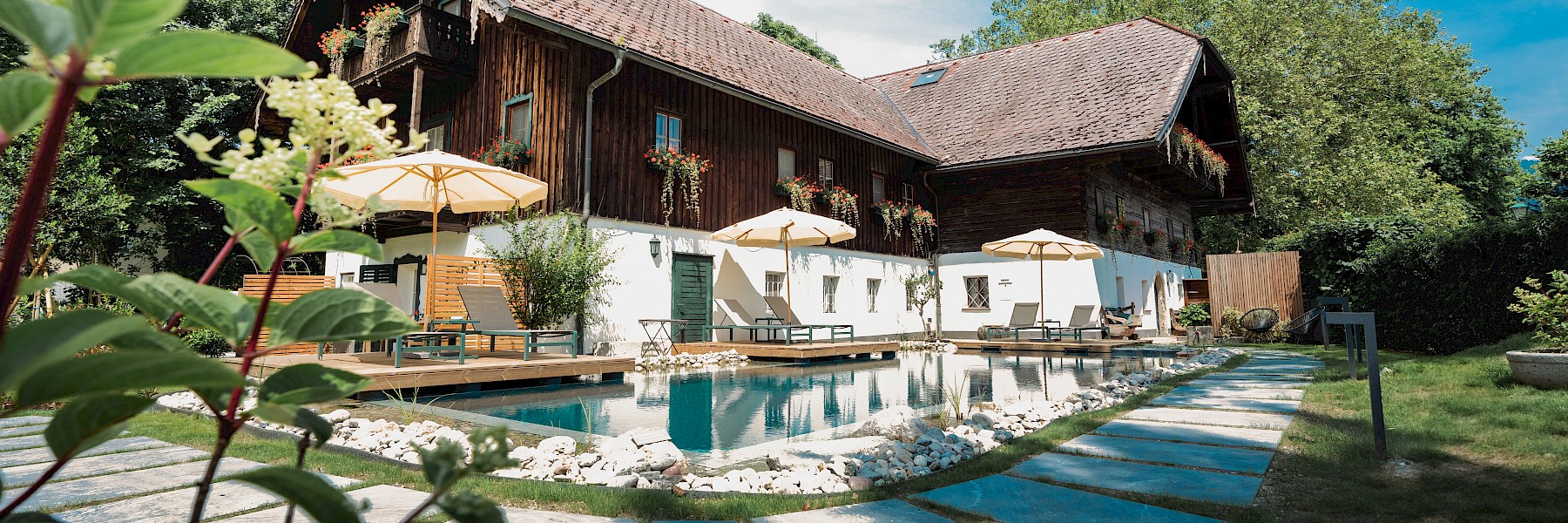 Das Bild zeigt einen idyllischen, gepflegten Garten mit einem Naturpool, umgeben von Liegestühlen und Sonnenschirmen vor einem charmanten, alten Landhaus mit Holzbalkon und blumengeschmückten Fenstern.